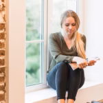 young-blonde-woman-holding-magazine-her-hand-sitting-window-sill_23-2147862856